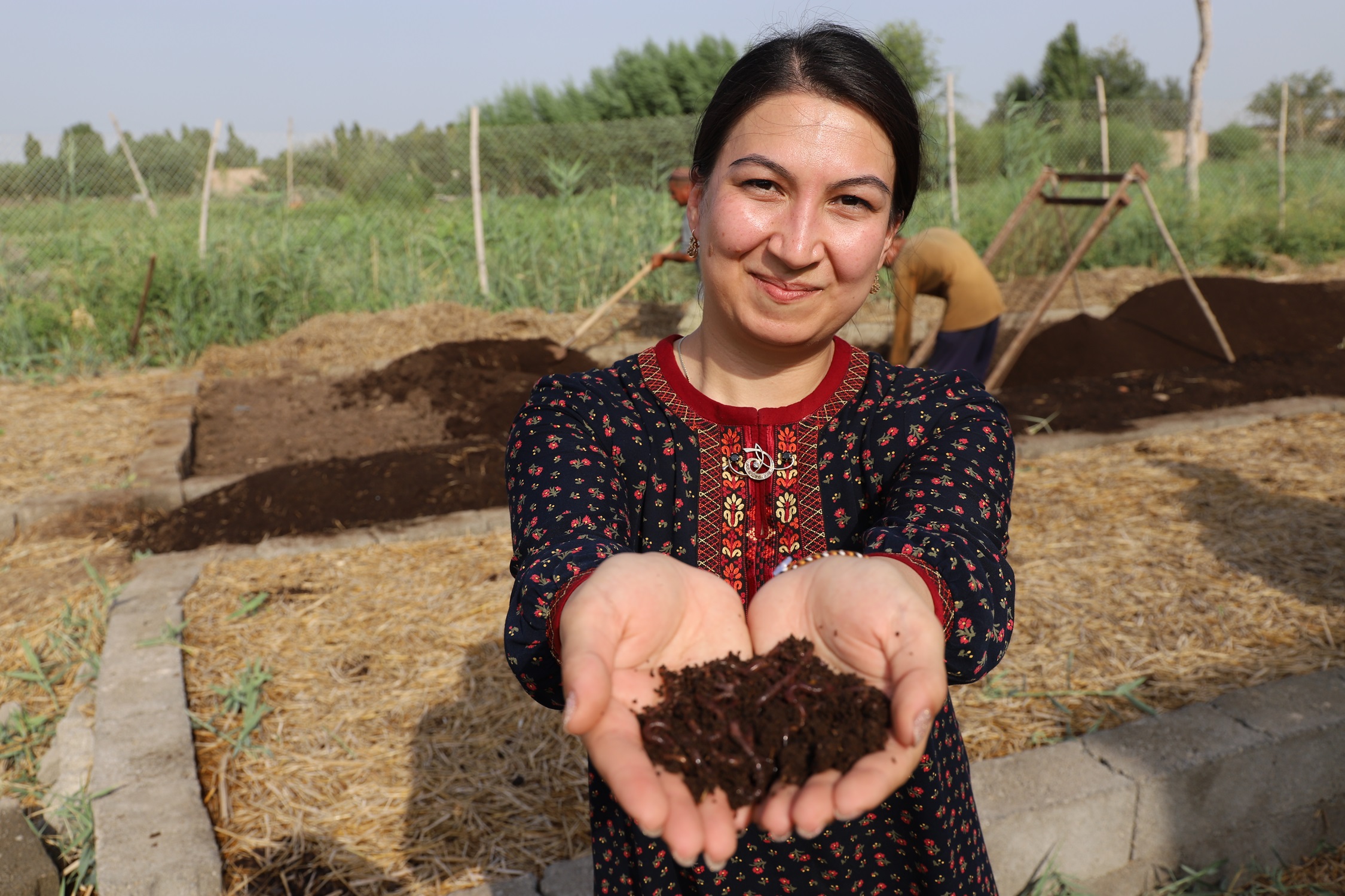A smiling woman holds soil in her hands, surrounded by a lush garden.